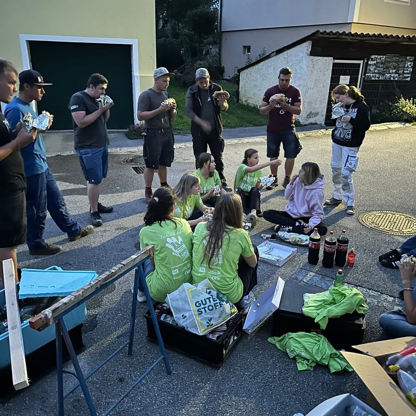 Eine Gruppe von Menschen in grünen T-Shirts versammelt sich um einen Tisch, isst Essen und sitzt auf dem Boden. Es gibt Kisten und eine Tasche auf dem Boden, und ein Mann mit Hut steht in der Nähe.