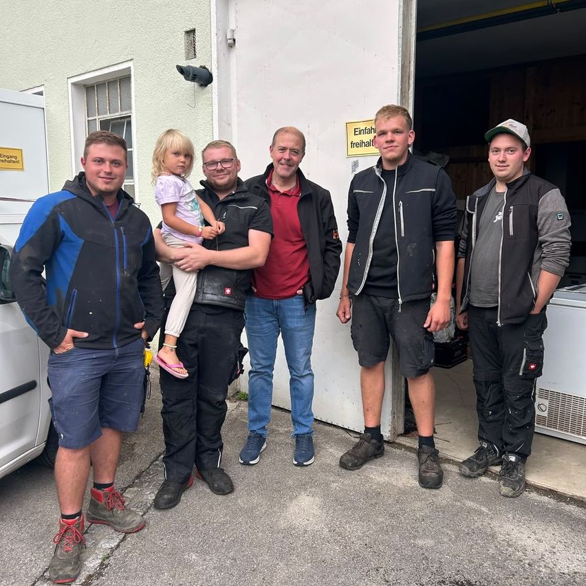 Fünf Männer und ein kleines Mädchen stehen vor einem Gebäude, zwei Männer halten das Mädchen, ein Mann trägt ein rotes Hemd und ein Schild steht 'Einfahrt freihalt'.