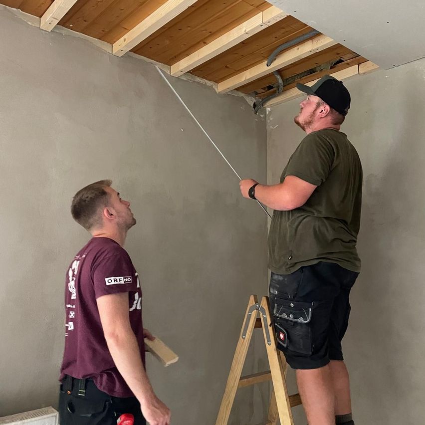 Zwei Männer arbeiten an der Decke eines Raumes, einer hält ein Werkzeug und der andere schaut nach oben. Der Raum hat Holzdeckenbalken und graue Wände.