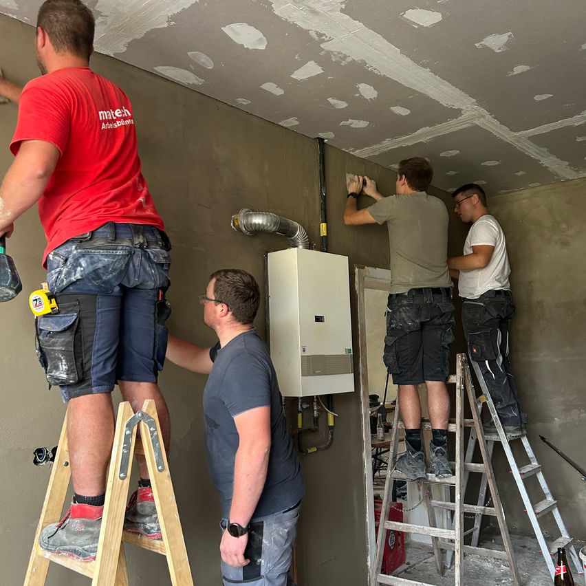 Vier Männer arbeiten auf einer Baustelle. Einer malt die Wand, ein anderer repariert etwas an der Wand und zwei stehen auf Leitern.