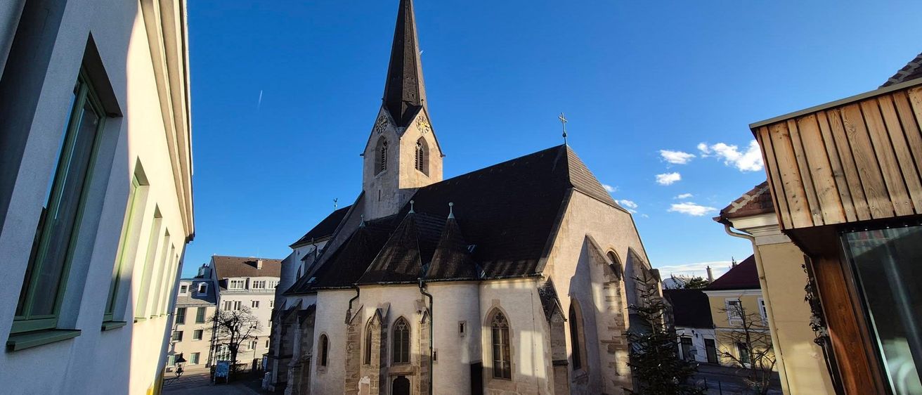 Eine gotische Kirche mit einem spitzen Turm und Uhren, vor einem klaren blauen Himmel, umgeben von Bäumen und Gebäuden.