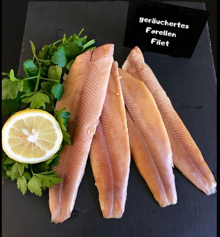 Ein Nahaufnahme von mehreren Fischfilets mit Petersilie und einer Zitronenscheibe auf einer schwarzen Oberfläche. Der Fisch ist als 'geräuchertes Forellen Filet' gekennzeichnet.