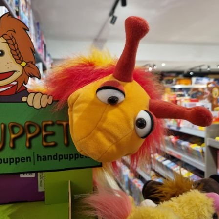 Ein gelbes und rotes Plüschtier mit großen Augen und einem Schild, auf dem PUPPET steht. Das Spielzeug ist in einem Geschäft mit anderen Spielzeugen auf Regalen.