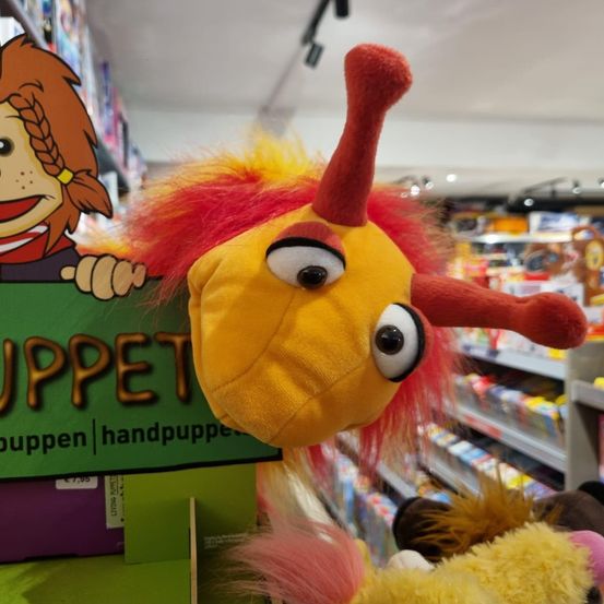 Ein gelbes und rotes Plüschtier mit großen Augen und einem Schild, auf dem PUPPET steht. Das Spielzeug ist in einem Geschäft mit anderen Spielzeugen auf Regalen.