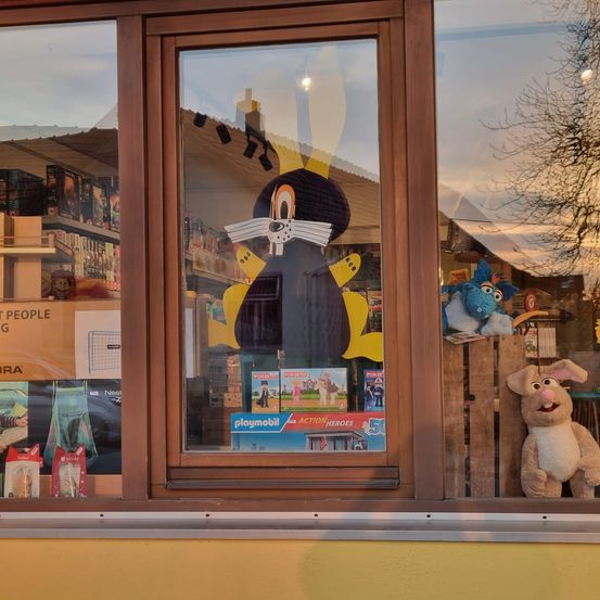 Ein Schaufenster eines Spielzeugladens zeigt eine große Zeichentrickfigur, Spielzeug und ein Plüschtier. Das Fenster ist von braunem Holz gerahmt.