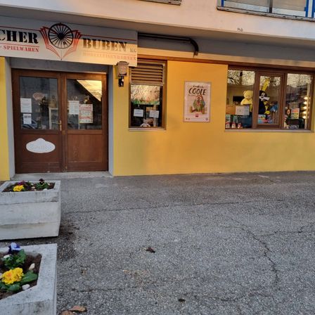 Ein gelbes Gebäude mit einer Holztür hat ein Schild, das 'Buben' mit einem Fahrrad darauf liest. Es gibt zwei Fenster mit Displays und einem Plakat. Das Gebäude hat Topfpflanzen und einen Betonsteig.