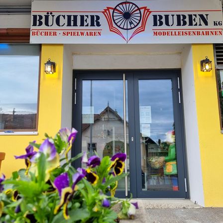 Ein gelbes Gebäude mit einem Schild, das Bucher Buben liest. Es hat eine Glastür mit einem Dummy und einer grünen Statue im Inneren. Im Vordergrund steht eine lila Blume.