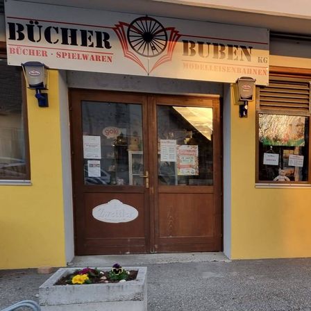 Die Fassade eines gelben Gebäudes hat ein Schild mit der Aufschrift 'BUBEN' und einem Fahrrad-Logo, zwei Türen und zwei Fenster. Zwei Betonpflanzgefäße mit Blumen davor.
