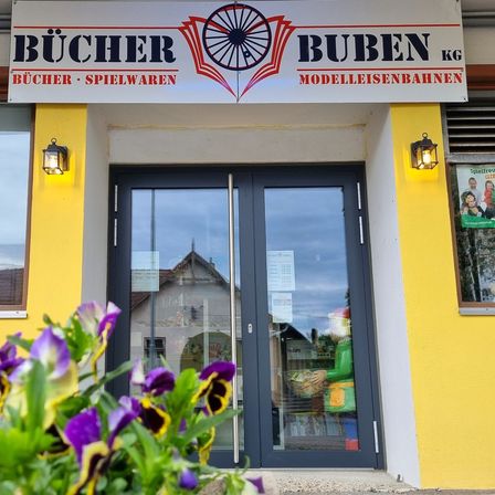 Die Ladenfront von Bucher Buben KG mit einem Schild über dem Eingang. Der Laden bietet Bücher und Spielzeug an und modellt Züge. Eine Lampe beleuchtet den Eingang, und eine Blumenpflanze steht davor.