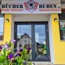 The storefront of Bucher Buben KG, featuring a sign above the entrance. The shop offers books and toys, and model trains. A lamp illuminates the entrance, and a flower plant is in front.
