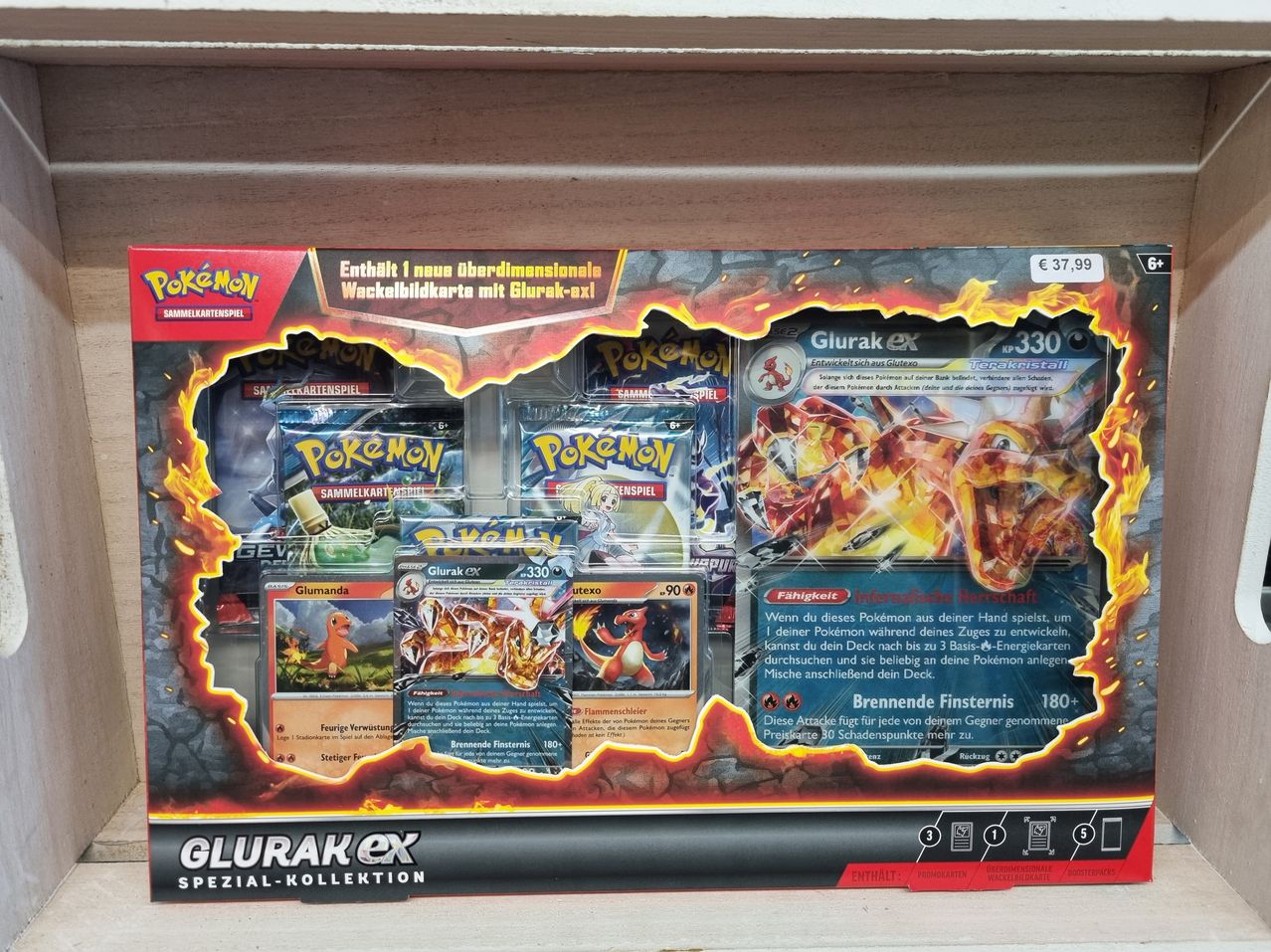 Eine Pokemon-Kartenbox zur Ausstellung. Sie enthält eine neue überdimensionale Karte mit Glurak-ex. Mehrere Karten und Packungen sind zu sehen, einige mit Feuereffekten. Preise sind auf der Box vermerkt.