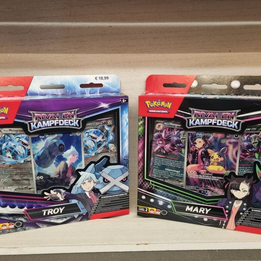 Zwei Packungen Pokemon-Karten sind auf einem Regal ausgestellt. Die linke Packung heißt 'Rivalenk Kampfdeck' mit der Figur Troy. Die rechte Packung heißt 'Mary' mit der Figur Mary.