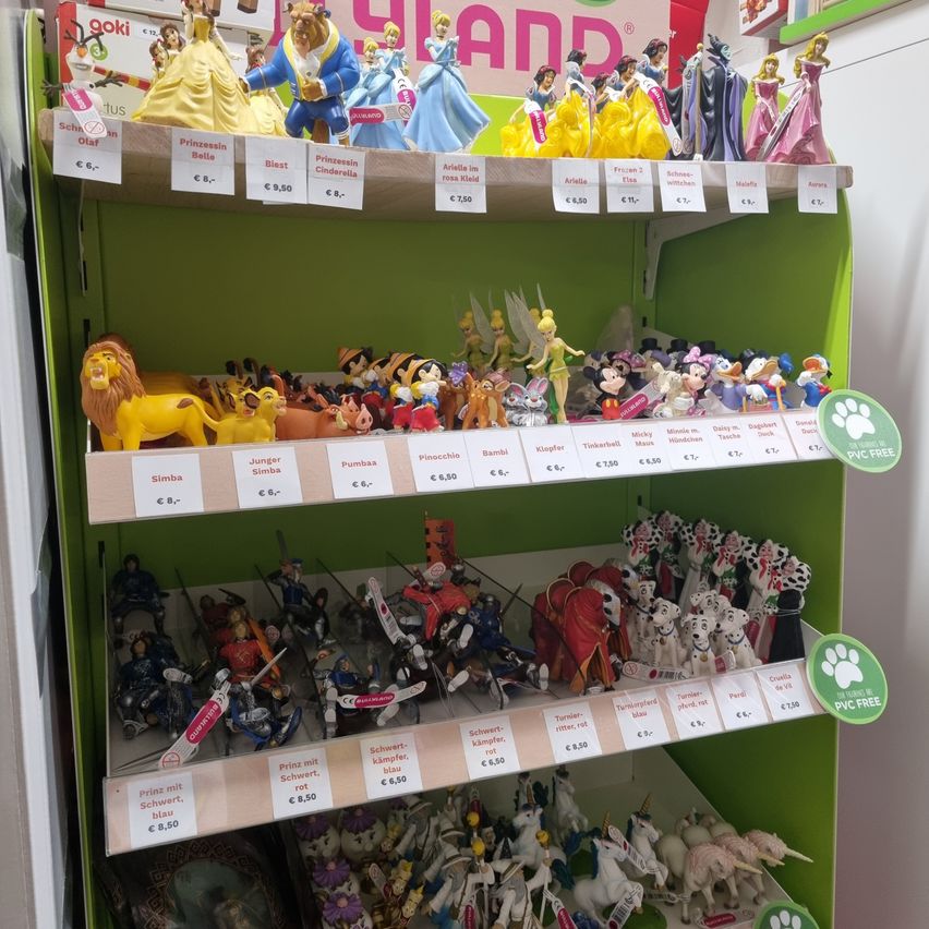 Ein grünes Regal mit mehreren Reihen von Spielzeug. Die oberste Reihe zeigt Disney-Prinzessinnen mit Preisschildern. Die zweite Reihe hat Micky Maus, Simba und andere Figuren. Die dritte Reihe hat Ritter und Einhörner.