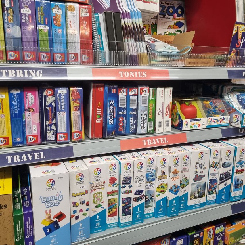 Ein Regal in einem Geschäft mit verschiedenen Brettspielen und Spielzeug. Das oberste Regal hat bunte Schachteln mit Text und Zahlen. Darunter befinden sich weitere Schachteln mit Bezeichnungen wie 'Domino', 'RoLife' und 'Sequence'. Das untere Regal ist mit 'Reise' beschriftet und hat eine Vielzahl von Schachteln.