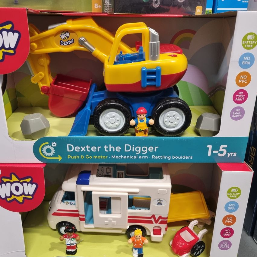 Ein verpacktes Spielzeugset mit einem gelben und roten Bagger, der den Markennamen WOW trägt. Der Bagger hat einen mechanischen Arm und klappernde Steine. Ein weißer Krankenwagen mit roten und weißen Streifen ist ebenfalls im Set enthalten.