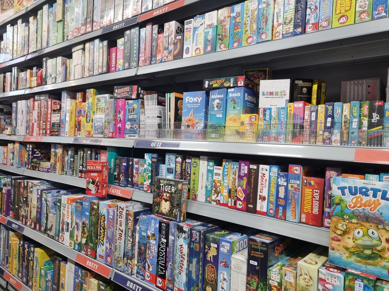 Ein Regal in einem Geschäft, gefüllt mit einer Vielzahl von Brettspielen und Büchern, die ordentlich in Reihen angeordnet sind und farbenfrohe und vielfältige Verpackungen zeigen.