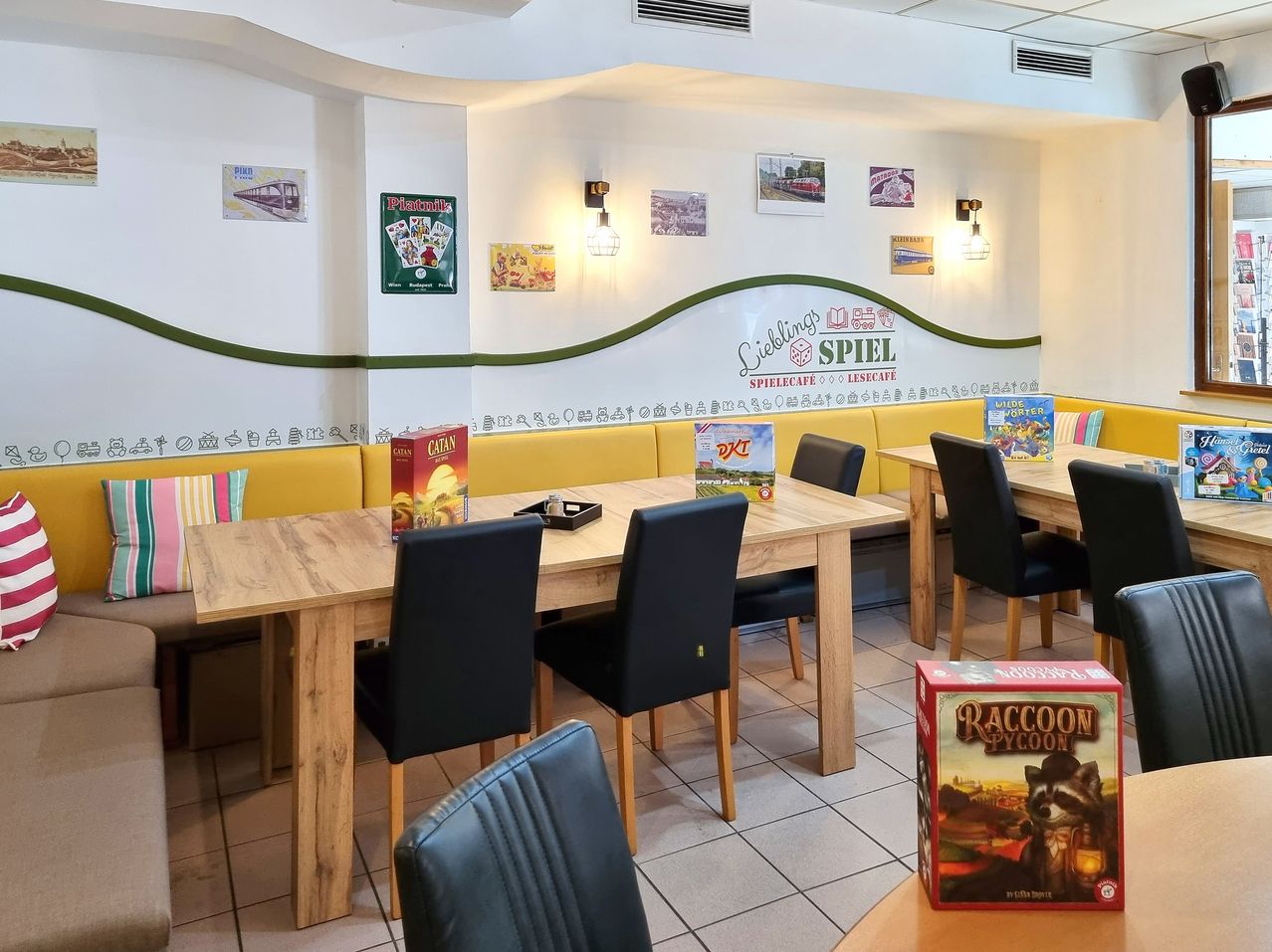 An indoor dining area with wooden tables and chairs. Several board games are on the tables. The wall has various decorations, including a green border, and a sign that reads Spielcafe.