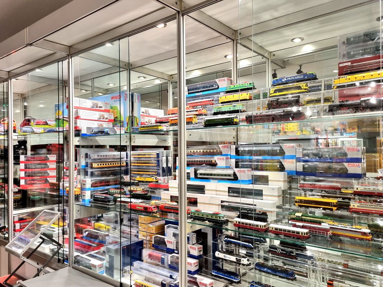 A glass display case filled with model trains and cars from different brands. The trains and cars are neatly arranged on multiple shelves. The trains are in various colors and styles.