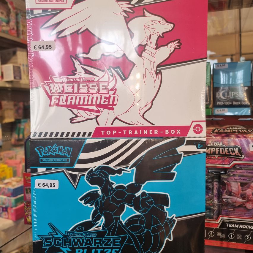 Two boxes of Pokemon cards on display in a store. The top box is white with a red border, featuring a drawing of a white dragon. Below it is a black box with a blue border, featuring a drawing of a black Pokemon. Both boxes are priced at 64,95 Euros.
