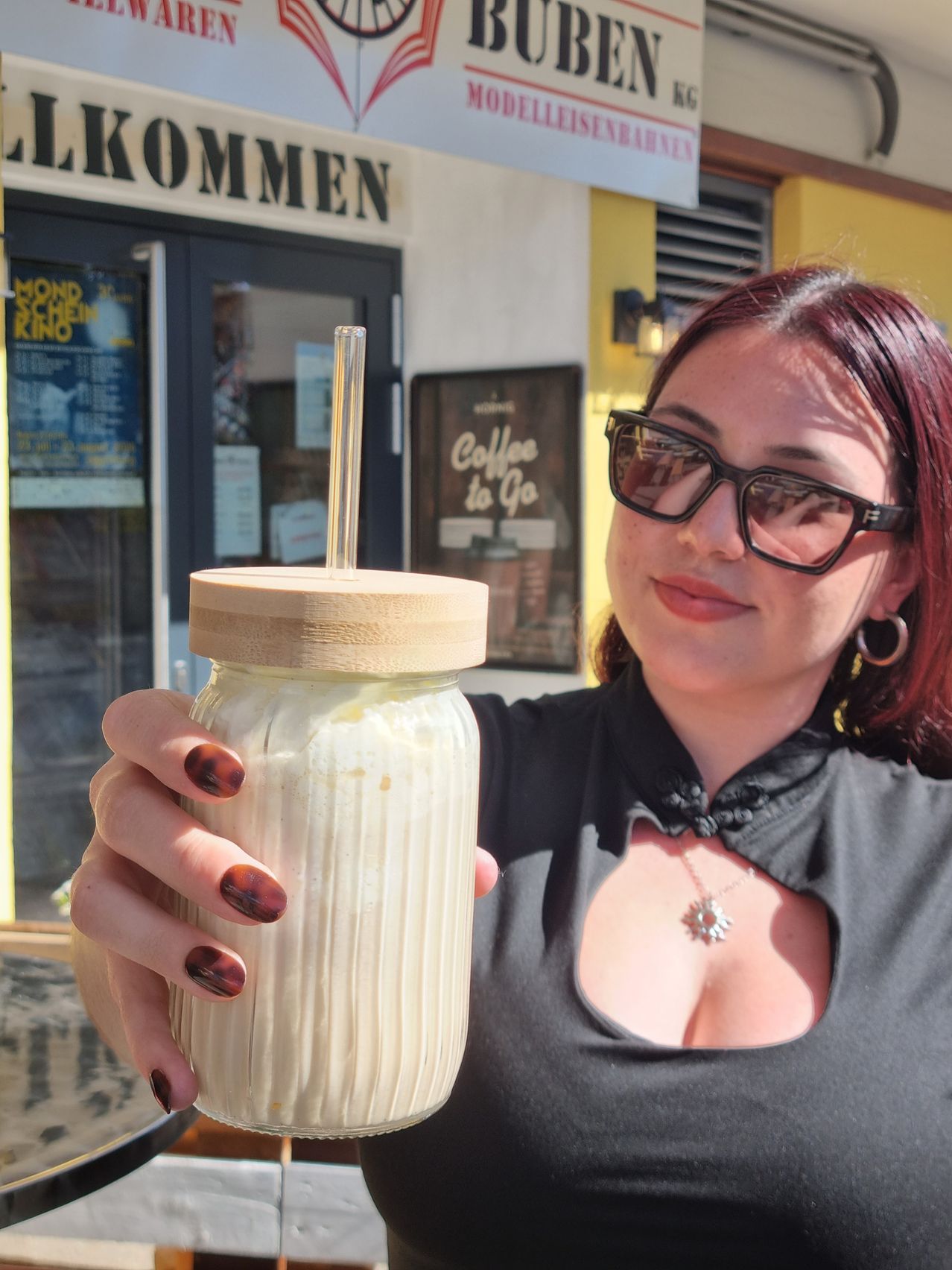 Eine Frau mit roten Haaren und einer Brille hält einen Glaskrug mit einem Holzdeckel und einem Strohhalm und lächelt vor einem Café.