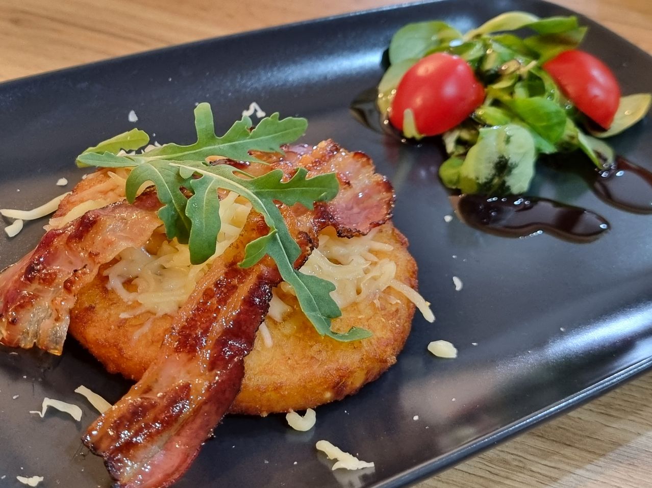 A golden fried potato cake with crispy bacon and cheese, garnished with arugula, on a black plate with a side salad.