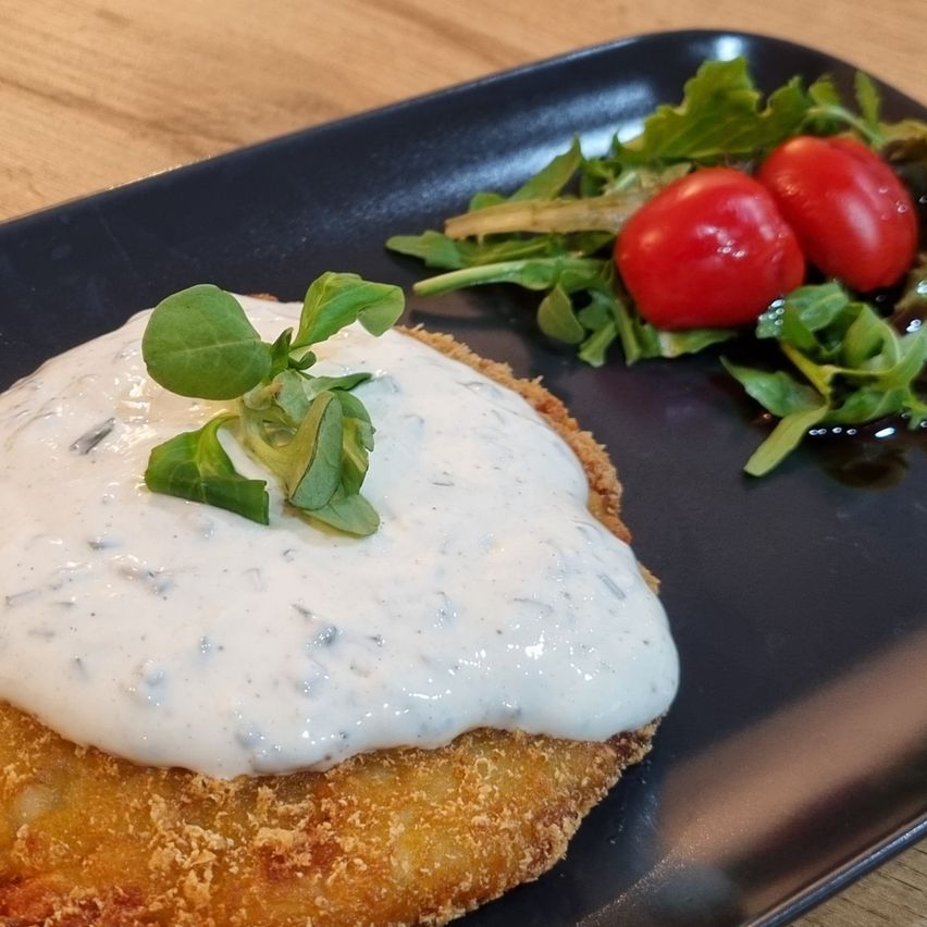 A fried fish cutlet topped with creamy sauce and garnished with green herbs, served on a black plate with a side of salad with cherry tomatoes.