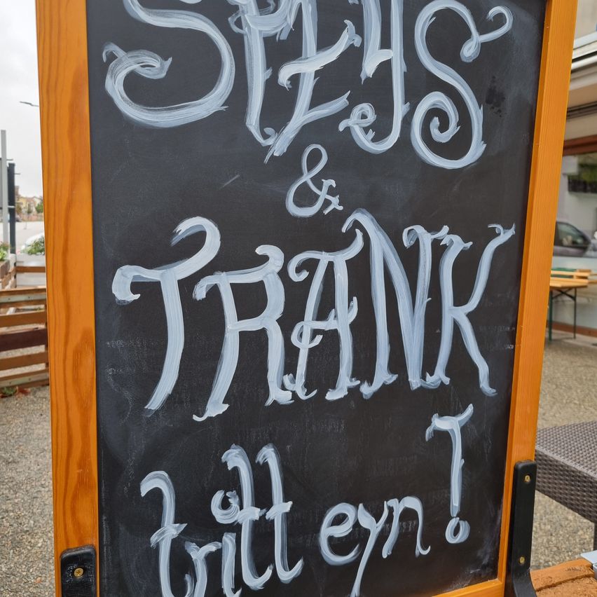 A chalkboard menu outside a restaurant features the words 'Spas & Frank' written in white chalk. Below it, the phrase 'bitten en?' is written in a smaller font. The background shows a blurry outdoor setting.