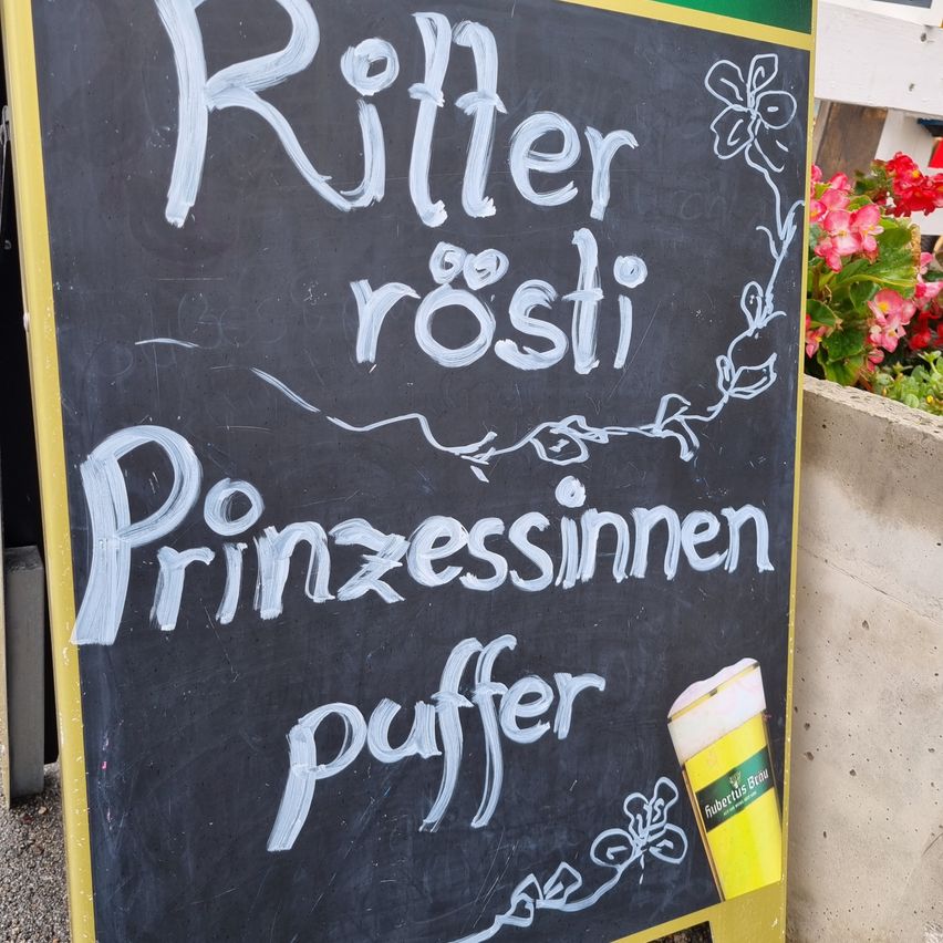 A blackboard with handwritten text in German, advertising 'Ritter rosi' and 'Prinzessinnen puffer'. A glass of beer is placed on the right side of the board.