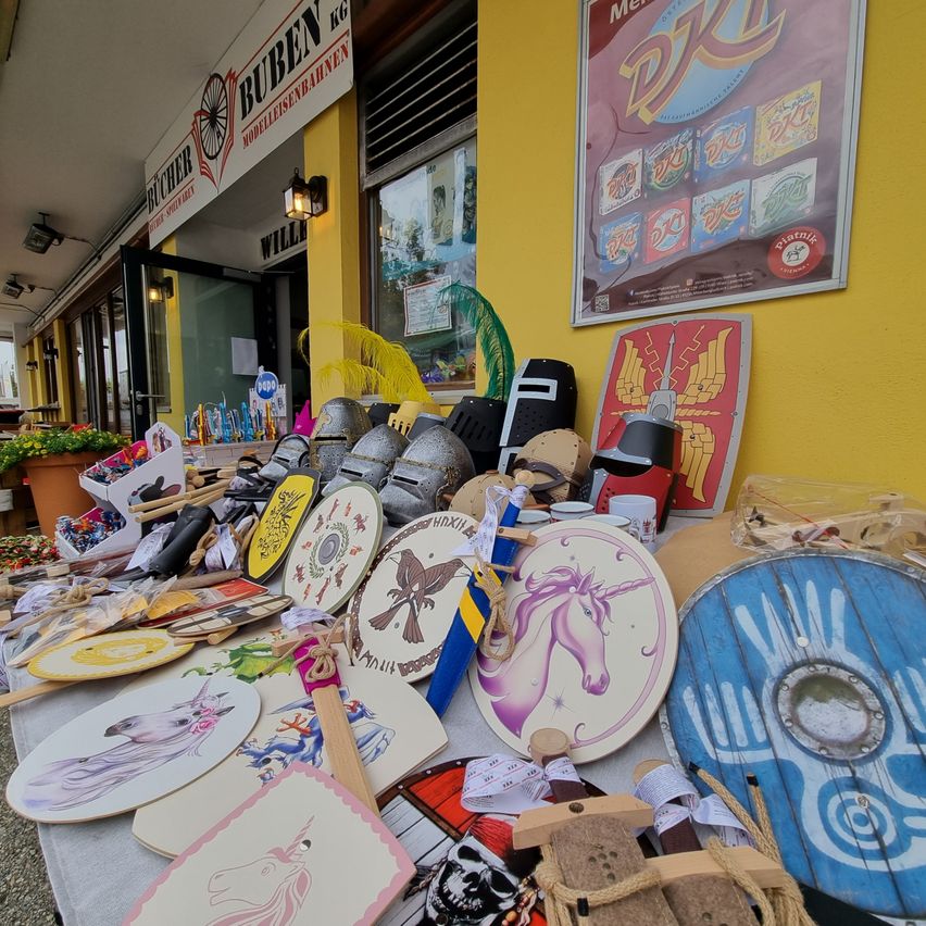A table displays various medieval-themed items, including shields, helmets, and swords, arranged in front of a store with a yellow wall and a sign reading 'BUBEN KG'.