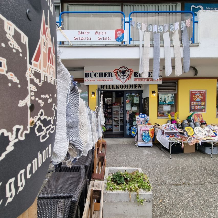 A storefront with a sign reading 'Bücher Spiele' is displayed. The entrance is open, and a table with various items is set up outside. A few chairs are placed in front of the store. A balcony with a railing and a banner is above the entrance.