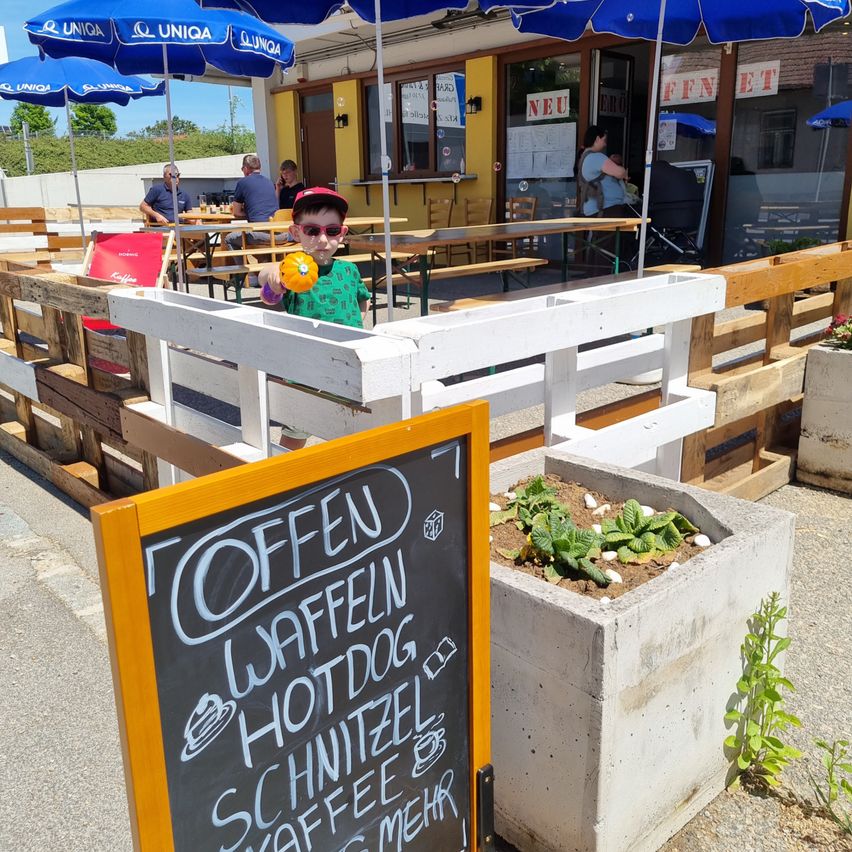 Ein junges Kind steht an einem Tisch vor einem Restaurant mit blauen Sonnenschirmen. Ein Kreidetafelmenü ist sichtbar.