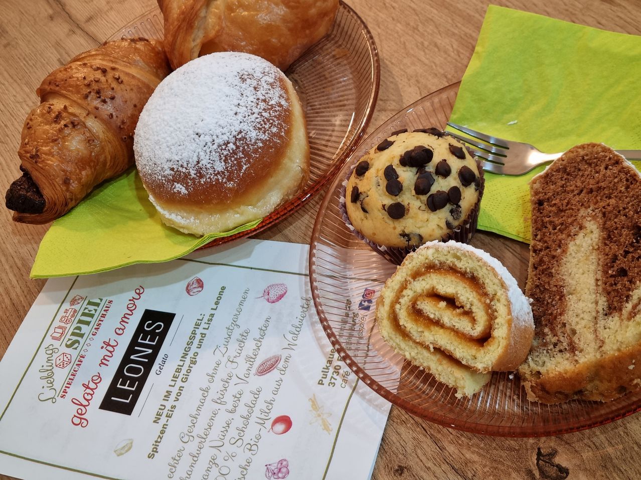 Two plates with various pastries, including a powdered donut, a chocolate chip muffin, and a rolled cake. A fork rests on a green napkin.