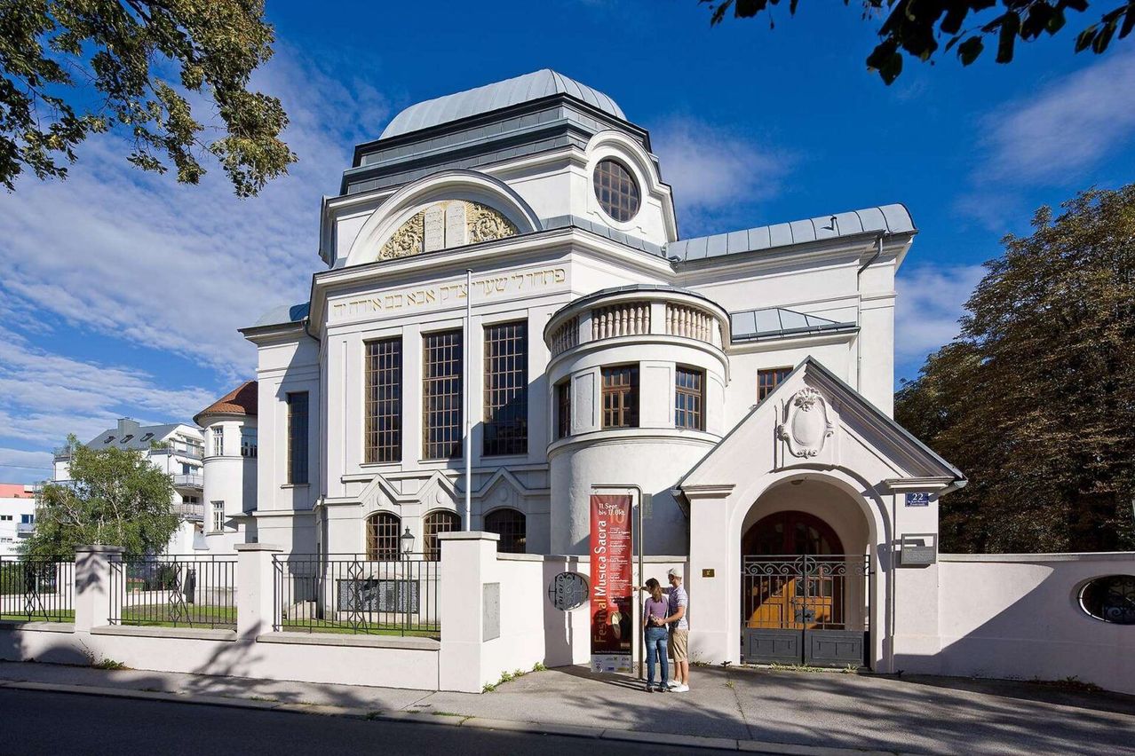 Ein weißes Gebäude mit einem Kuppeldach und Rundbogenfenstern. Zwei Personen stehen davor. Das Gebäude hat ein rotes Schild mit weißem Text.