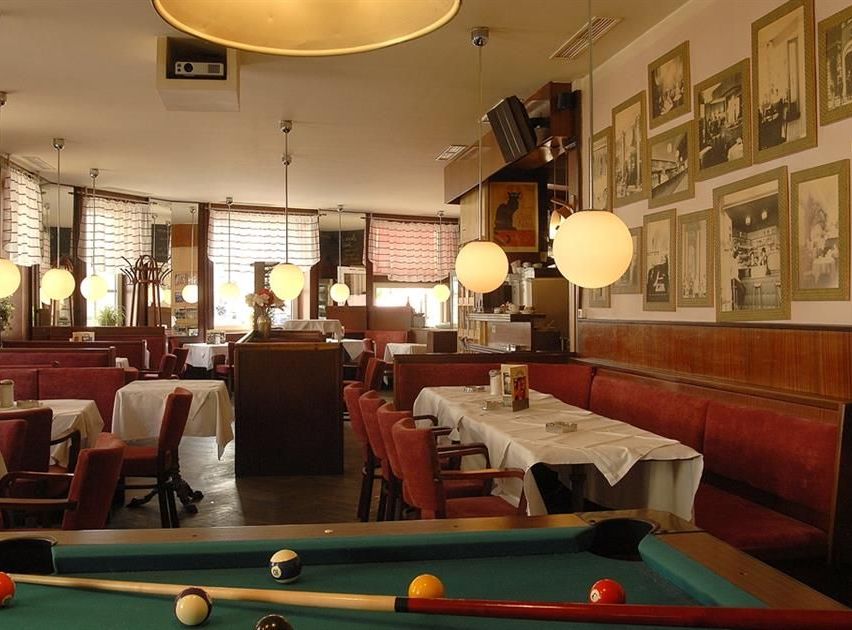 Ein Restaurant mit Billardtisch im Vordergrund, Tische mit weißen Tischdecken, Stühlen und einer hölzernen Theke mit Monitor. Eine Projektorlampe an der Decke und viele Bilderrahmen an der Wand.