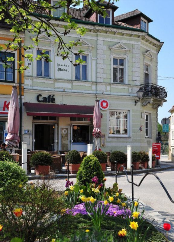 Ein Café mit einem roten Vordach und einem Balkon befindet sich an einer Straße mit blühenden Pflanzen und Topfpflanzen.