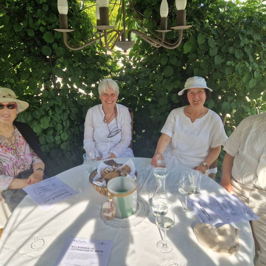 Vier Personen sitzen um einen Tisch in einem Garten. Zwei Frauen tragen Hüte und weiße Blusen, während ein Mann eine Uhr trägt. Auf dem Tisch stehen ein Korb mit Keksen, Weingläser und Papiere. Über ihnen hängt eine Kronleuchter.