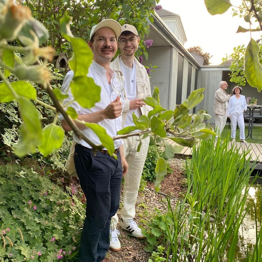 Zwei Männer stehen in einem Garten, einer zeigt den Daumen nach oben. Hinter ihnen steht ein Haus mit einer kleinen Veranda. Im Hintergrund stehen zwei ältere Menschen. Es gibt einen Teich mit hohem Gras und Grünpflanzen.