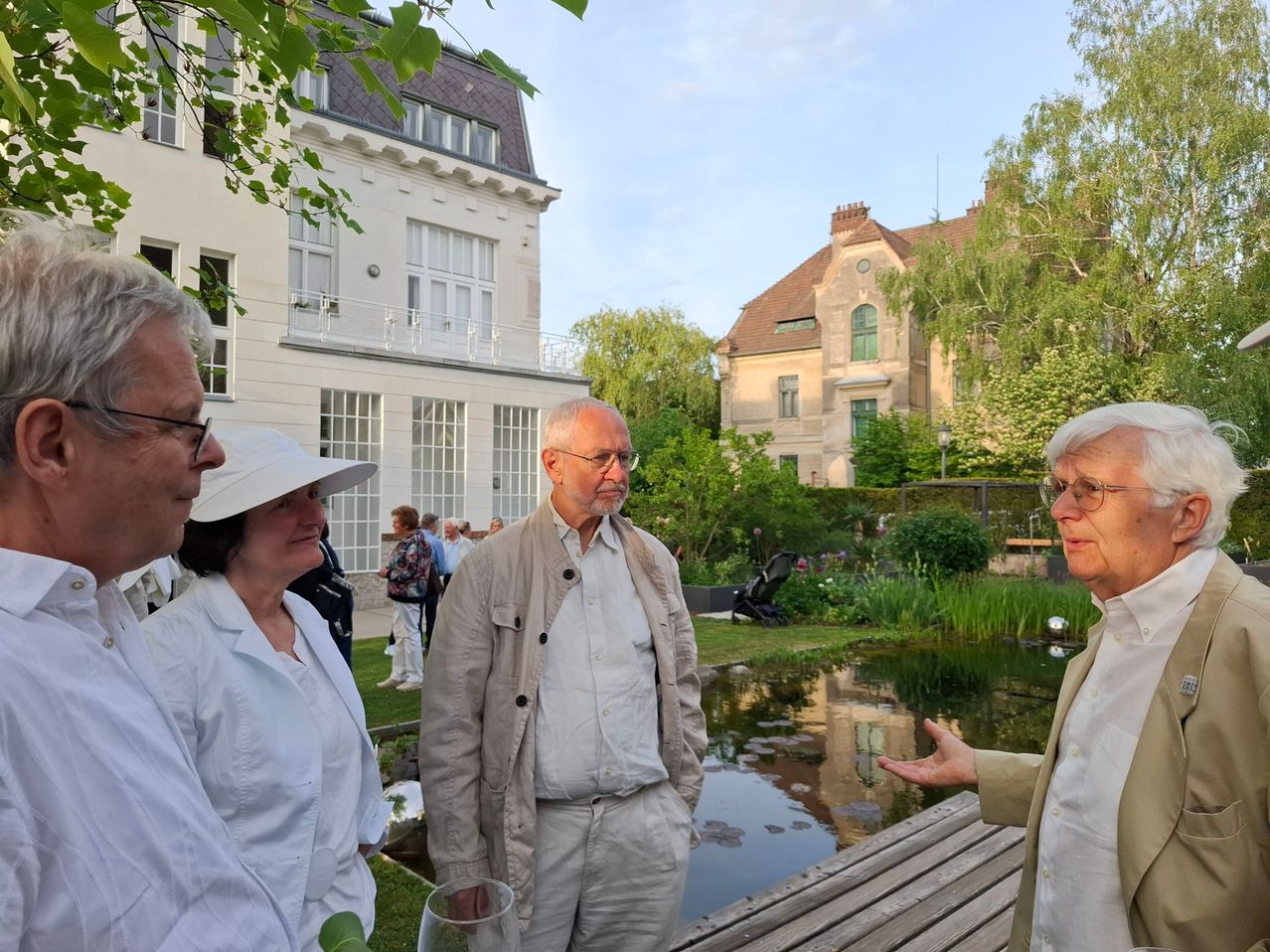 Eine Gruppe von Menschen steht vor einem Haus mit einem Teich und Garten. Der Mann in der Mitte trägt eine Brille und eine beige Jacke.
