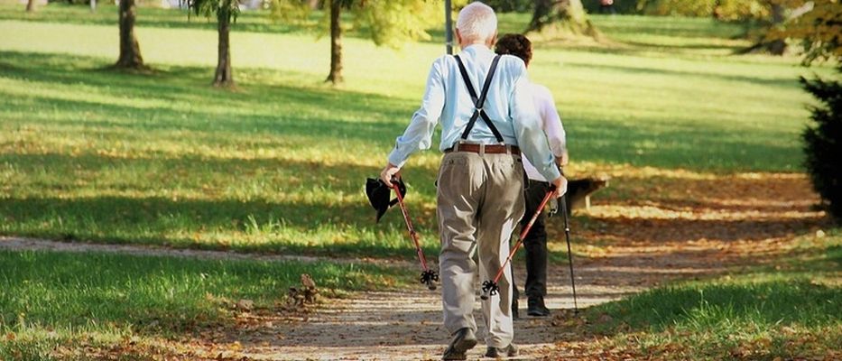 Ein älterer Mann mit Hosenträger geht mit einem Stock begleitet von einer Frau auf einem Grasweg.
