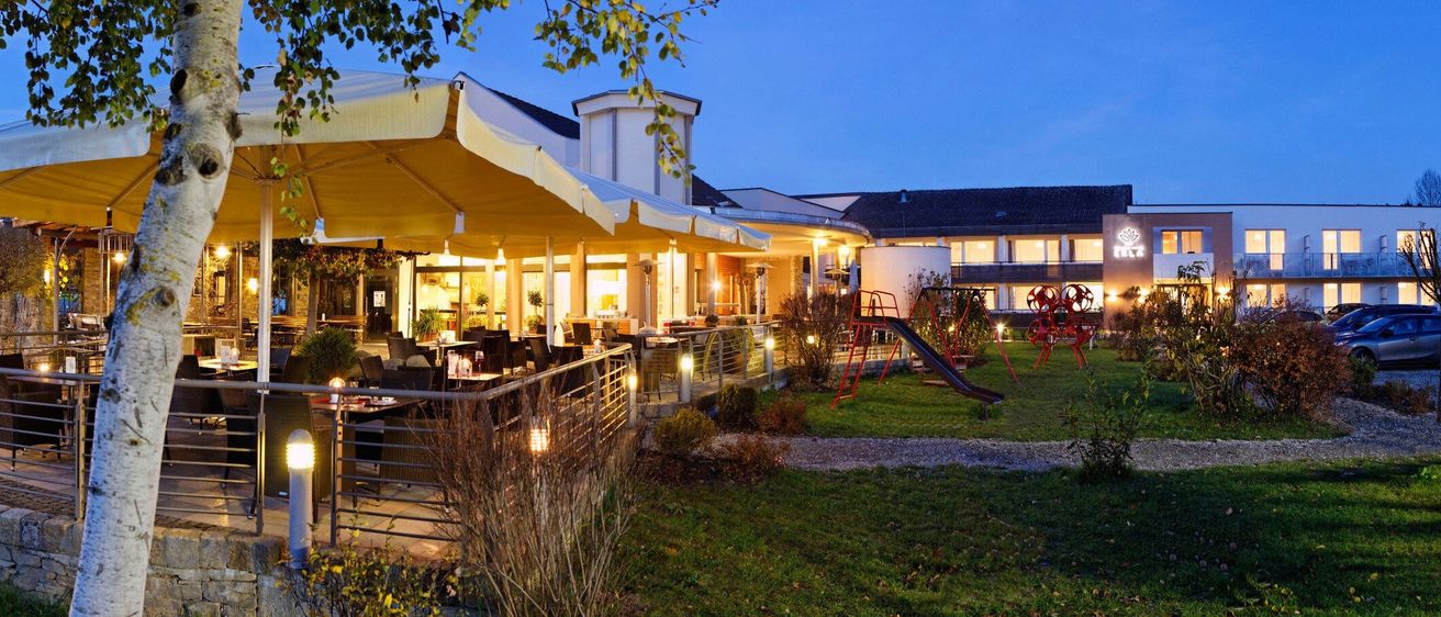 Ein Restaurant im Freien in einem Hotelkomplex am Abend mit einem großen Regenschirm und Tischen, Stühlen und Lampen.