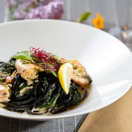 Ein weißer Teller mit einem Gericht aus schwarzen Nudeln, Garnelen und Garnierung.