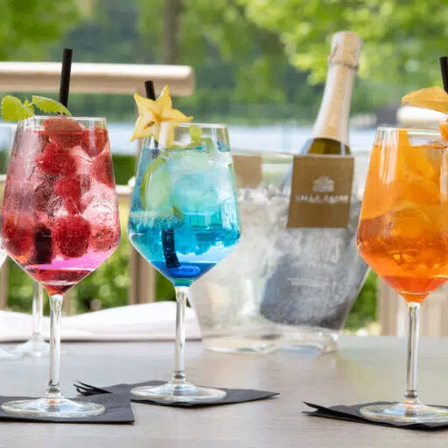 Drei farbenfrohe Cocktails mit dekorativen Garnierungen stehen auf einem Tisch. Im Hintergrund befinden sich zwei leere Weingläser und eine Flasche Champagner.