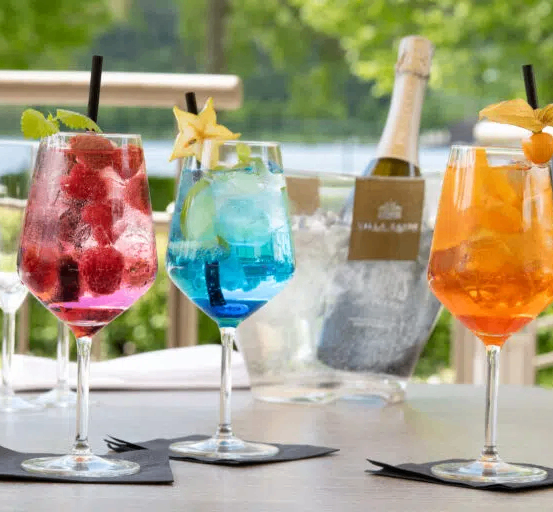Drei farbenfrohe Cocktails mit dekorativen Garnierungen stehen auf einem Tisch. Im Hintergrund befinden sich zwei leere Weingläser und eine Flasche Champagner.