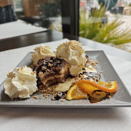 Ein Teller mit Dessert zeigt einen Kuchen mit Schlagsahne, Schokoladensirup und Orangenscheiben auf einem Tisch.