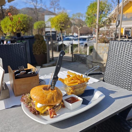Ein Burger mit Speck und Käse auf einem weißen Teller, begleitet von Pommes, einer kleinen Schüssel Ketchup und Besteck auf einem Tisch in einem Außenrestaurant.