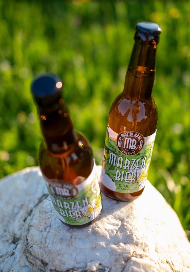 Zwei braune Flaschen Marzen Bier stehen auf einem Felsen. Eine Flasche hat ein dunkelgrünes Etikett, die andere ein hellgrünes. Der Hintergrund ist ein verschwommener grüner Rasen.