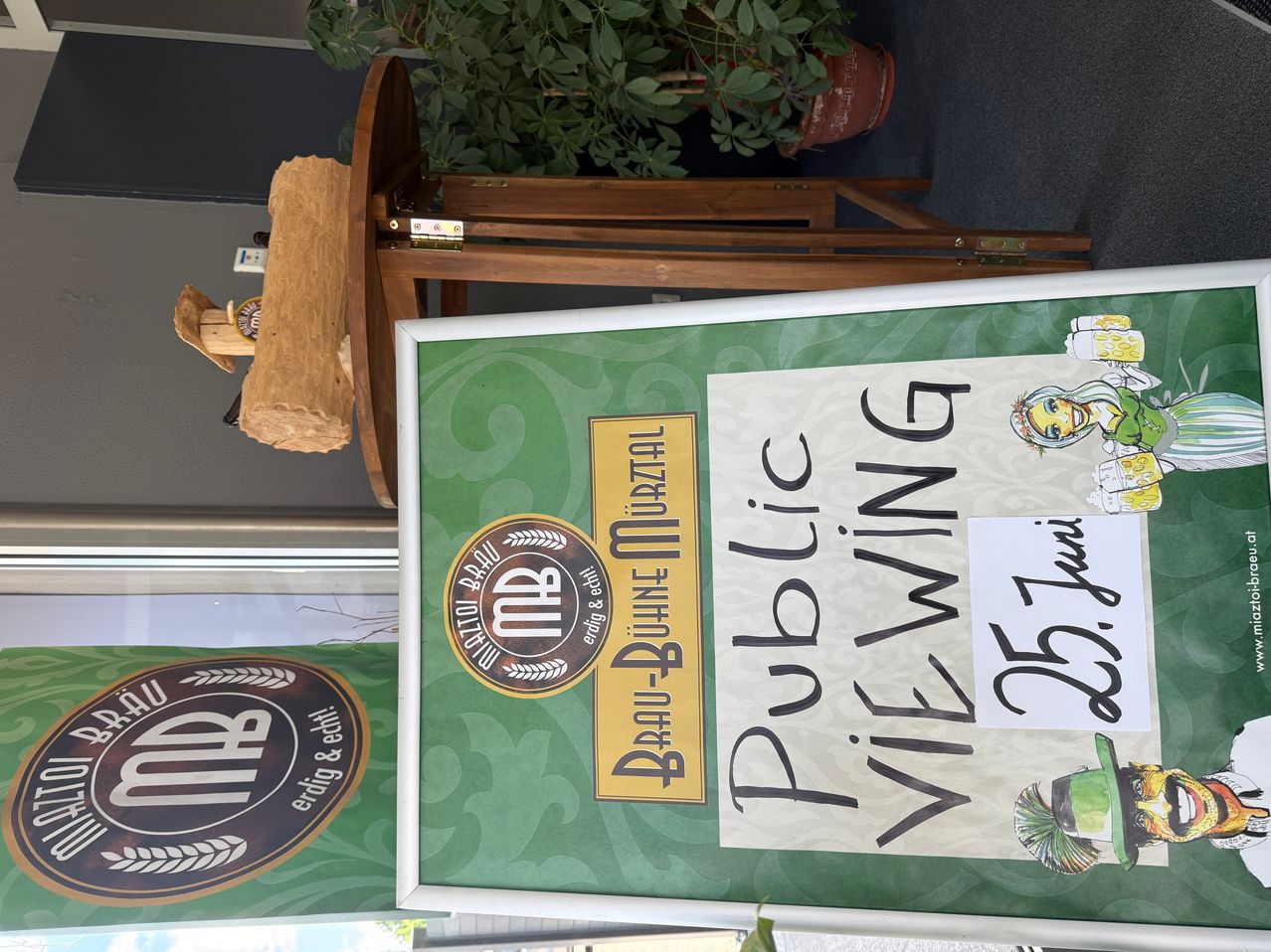 Ein gerahmter Schild mit der Aufschrift 'Public Viewing'. Darunter befindet sich ein gelbes Etikett mit 'Braeu-Delle-Muerzthal'. Auf der linken Seite ist ein Logo mit 'MB'. Eine Pflanze befindet sich hinter dem Schild.