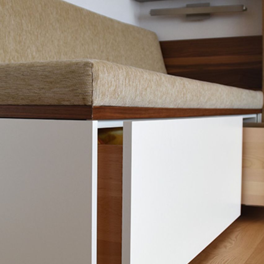 Eine Nahaufnahme einer weißen Bank mit beigefarbenen Kissen und einem Holzrahmen, mit einer Schublade darunter. Die Bank steht an einer Holzwand mit einer montierten Leuchte.