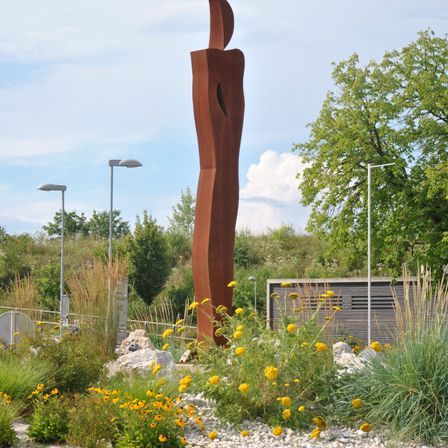 Eine große braune Skulptur steht inmitten eines Gartens mit gelben Blumen, Steinen und hohen Gräsern. Dahinter sind drei Straßenlampen zu sehen. Bäume und ein Gebäude im Hintergrund.