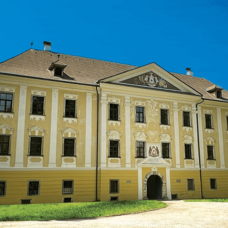 Ein großes gelbes Gebäude mit vielen Fenstern und einem braunen Dach steht auf einem Rasen unter einem klaren blauen Himmel.
